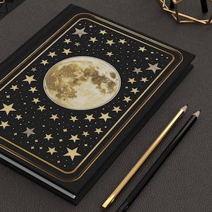 Peut inclure: Journal noir avec une bordure dorée, un motif de lune et d'étoiles. La couverture présente une grande illustration de lune entourée d'étoiles dorées. Le texte indique "150 pages lignées" et "Finition mate".