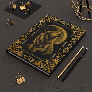 May include: Black journal with a gold illustration of a howling wolf against a moon. The cover features a gold ornate border. Gold pencils and binder clips are on a dark gray surface.