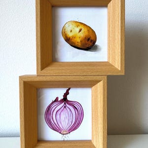 Peut inclure: Deux cadres carrés en bois clair empilés verticalement. Le cadre supérieur contient une peinture d'une pomme de terre brune. Le cadre inférieur contient une peinture d'un oignon rouge coupé en deux montrant sa structure interne violette et blanche.