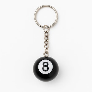 Puede incluir: Un llavero de bola ocho en blanco y negro. El llavero tiene un anillo y una cadena de metal plateado. La bola ocho es de color negro brillante con el número ocho blanco en un círculo blanco.