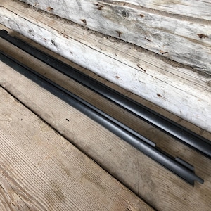 May include: Two long, black, rectangular pieces of hardware, possibly for a window or door, are positioned diagonally on a weathered wooden surface. The wood has a natural, aged appearance with visible grain and knots.