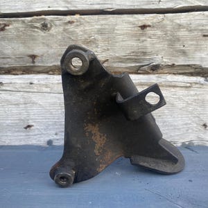 Puede incluir: Un soporte automotriz de metal oscuro y desgastado con forma triangular y tres puntos de montaje distintos, que incluyen dos orificios circulares y una lengüeta saliente con un orificio, mostrando signos de oxidación y desgaste en toda la superficie.