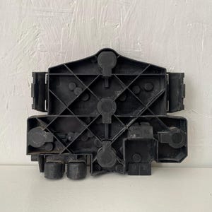 May include: A black plastic automotive component featuring a complex structural grid pattern with multiple circular mounting points, rectangular side tabs, and two cylindrical protrusions at the bottom edge, designed for secure mechanical assembly.