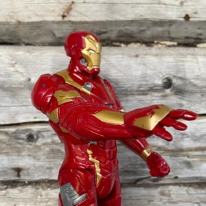 May include: A red and gold Iron Man action figure, posed with an outstretched arm. The figure has a red and gold metallic finish, with intricate details on the armor. The background is a weathered wooden surface.