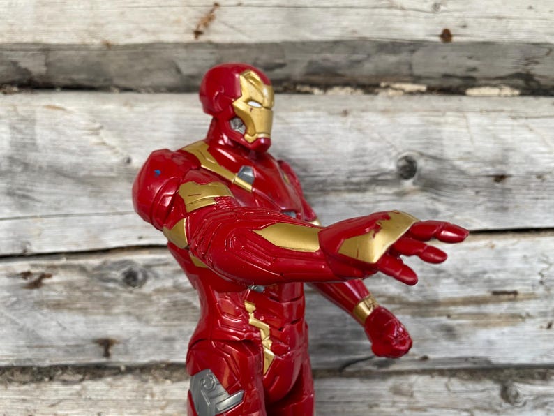 May include: A red and gold Iron Man action figure, posed with an outstretched arm. The figure has a red and gold metallic finish, with intricate details on the armor. The background is a weathered wooden surface.