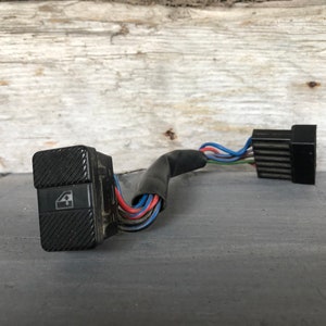 Puede incluir: Un interruptor de ventana de coche negro con una superficie texturizada y el número 4. Está conectado a un mazo de cables con cables de colores. Este interruptor probablemente sirve para controlar las ventanillas eléctricas de un vehículo.
