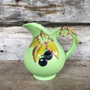Puede incluir: Una jarra de cerámica verde claro con un cuerpo redondeado y un asa curva. La jarra está decorada con diseños florales y de mora en relieve en tonos marrón, amarillo y negro. La jarra tiene un pico para verter.