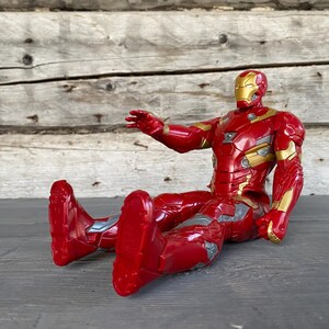 May include: A red and gold Iron Man action figure in a seated position. The figure has a metallic appearance with gold accents on the chest, arms, and helmet. The figure is approximately 6 inches tall.