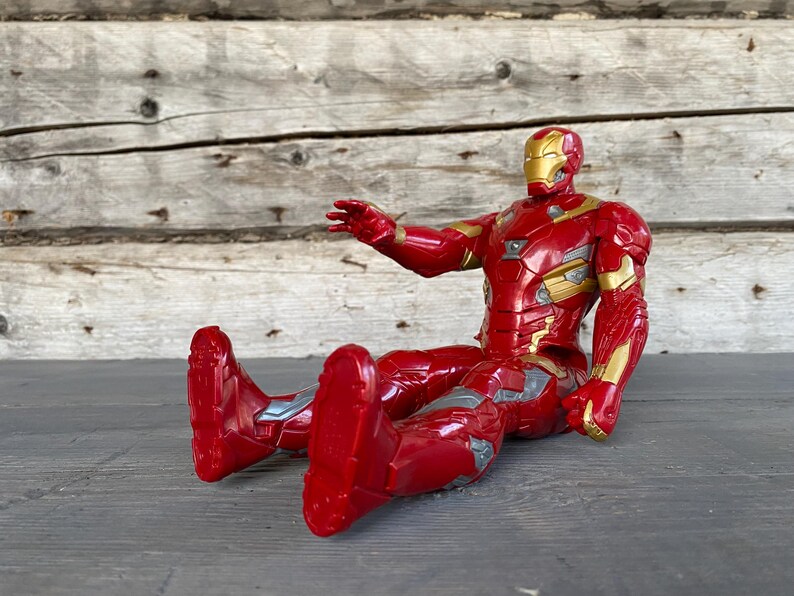May include: A red and gold Iron Man action figure in a seated position. The figure has a metallic appearance with gold accents on the chest, arms, and helmet. The figure is approximately 6 inches tall.