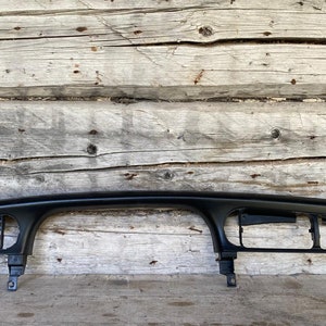 Puede incluir: Un embellecedor negro para el salpicadero de un coche. El embellecedor tiene un diseño curvo con aberturas rectangulares y pestañas de montaje. El fondo son tablas de madera desgastadas.