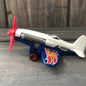 May include: A toy airplane with a white fuselage, blue wings, and a red propeller. The plane has a silver engine detail and a flame design on the wing. The toy is on a wooden surface.