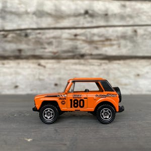 May include: Orange and black toy SUV with the number 180 on the side. The toy car has black wheels and a spare tire on the back. The words "4x4", "MP" and "Bushwacker" are also visible.