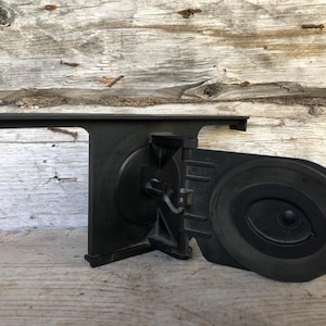 Può includere: Un componente per auto in plastica nera, possibilmente un altoparlante o una staffa di montaggio, con un elemento circolare e una sezione rettangolare. Il pezzo è posto su uno sfondo di legno invecchiato.