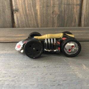 May include: A black and silver toy car with a gold horn-like structure on top. The wheels are black with a skull design. Red accents are visible on the car's body.