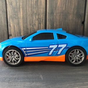 May include: A blue toy race car with orange accents and the number 77 on the side. The car has silver wheels and a spoiler. The toy car is set against a wooden background.