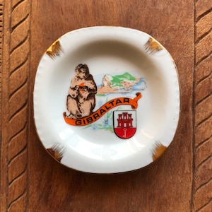 Puede incluir: Un cenicero pequeño y blanco con borde dorado. El cenicero presenta una colorida ilustración de un mono, un paisaje y la palabra "GIBRALTAR" en una pancarta naranja. También se muestra un escudo de armas.