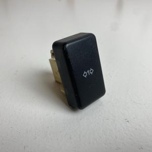 May include: A rectangular black plastic electrical switch featuring a white icon of the number one flanked by two horizontal arrows pointing in opposite directions. The switch has a smooth, matte finish and a beige plastic base component.