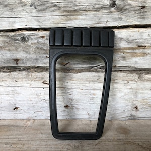 May include: A black plastic car console trim piece. The trim has a rectangular opening and a series of raised rectangular sections at the top. The piece is set against a weathered wooden background.