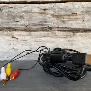 May include: Black AV cable with yellow, red, and white RCA connectors. The cable is coiled and has a black connector on one end. The background is a weathered wooden surface.