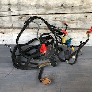 May include: A tangled bundle of black automotive wiring harnesses with various colored connectors, including red, yellow, and green. The wires are against a weathered, light-colored wooden background.