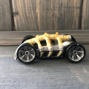 May include: A toy car with a skeleton-like design. The car features a yellow bone structure on top of a black chassis, with silver-colored wheels. The wheels have a detailed spoke design. The car is set against a wooden background.