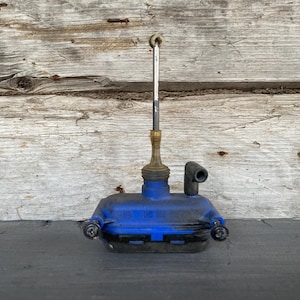 Puede incluir: Una aceitera azul y negra vintage con un pico plateado y de latón. La lata tiene una base rectangular y un pico curvo. El fondo es una superficie de madera desgastada.