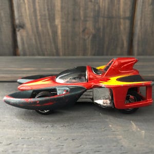 May include: A red and black toy car with yellow flame details. The futuristic vehicle has a black cockpit and three wheels. The toy car is on a gray surface with a wooden background.