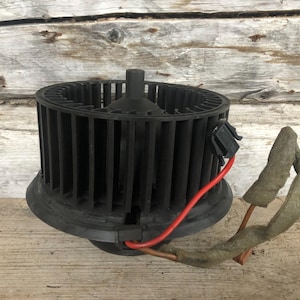 Puede incluir: Un motor de ventilador automotriz negro con un diseño cilíndrico y aletas verticales. Un cable rojo y un conector negro están unidos. El motor está sobre una superficie de madera.