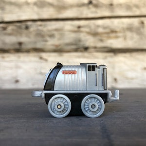 May include: A silver toy train with black detailing, featuring the name "SPENCER" in a red rectangle. The toy train has two sets of wheels and is set against a wooden backdrop.