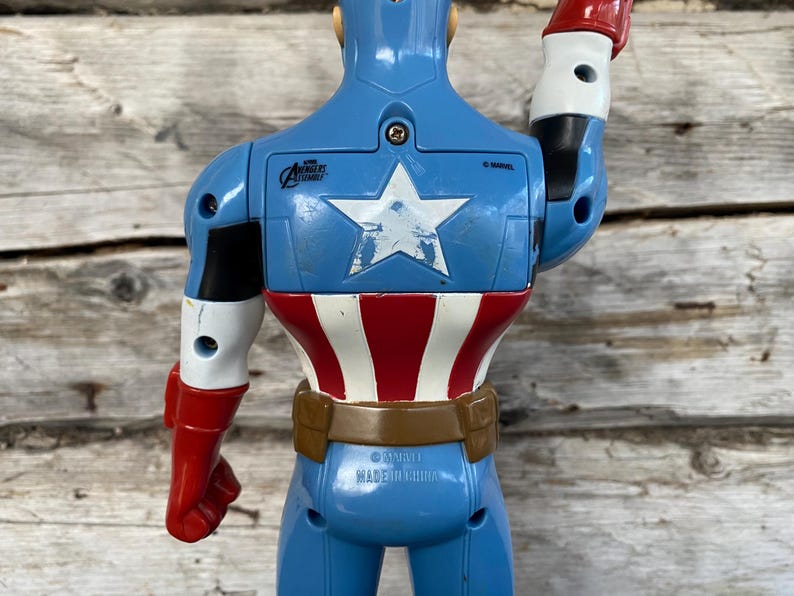 May include: A blue Captain America action figure, viewed from the back. The figure has a white star on the chest, red and white stripes on the torso, and red gloves. The figure is made in China.