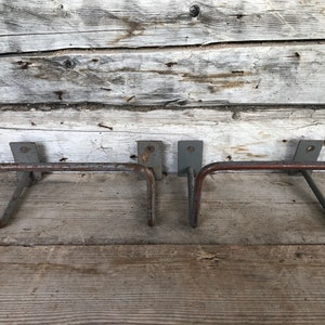May include: Two vintage metal brackets with a rusty finish. Each bracket has a rectangular mounting plate and a curved, handle-like bar. The brackets are mounted on a weathered wooden surface, suggesting an antique or industrial aesthetic.