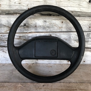 Puede incluir: Un volante negro con un cubo central rectangular con el logotipo de Volkswagen. El volante es circular con un agarre grueso y texturizado. El fondo es de madera clara desgastada.