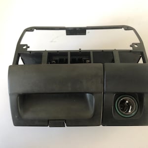 May include: A black plastic car dashboard component with a rectangular opening, a small compartment, and a circular opening. The component has a textured surface and is likely a replacement part.