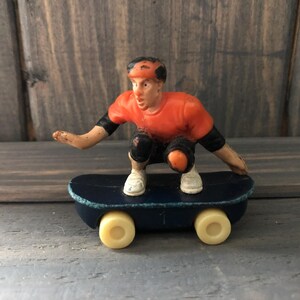 Può includere: Figurina di skateboarder in miniatura con maglietta arancione, gomitiere e ginocchiere nere e scarpe bianche, accovacciata su uno skateboard blu navy con ruote gialle. La figurina ha un casco con dettagli arancioni e neri.