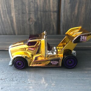 May include: A gold-colored toy tow truck with purple wheels. The truck has a shiny finish and features the text "24K Service" and "Mother Load". The toy is set against a wooden background.