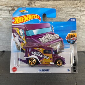 May include: A Hot Wheels die-cast toy car in a package. The car is a purple and gold "Mailed It!" delivery truck with gold wheels. The packaging includes the Hot Wheels logo and the text "MAILED IT!"