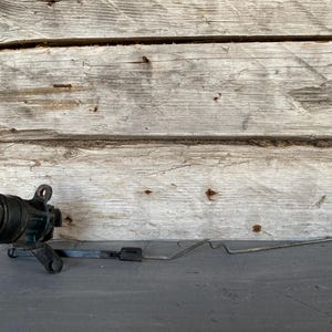 Puede incluir: Un cilindro de cerradura de puerta de automóvil negro con un ojo de cerradura, unido a una varilla metálica y un soporte negro. El conjunto se encuentra sobre un fondo de madera desgastada.