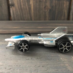 May include: A silver toy race car with black wheels and blue accents. The car has the number "909" printed on the side and rear, along with other blue graphics. The car is a miniature replica of a race car.