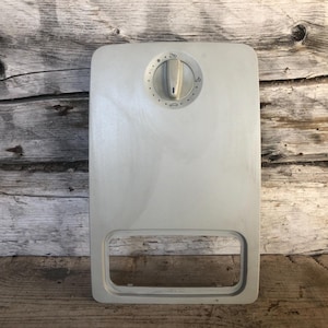 May include: A light gray appliance control panel with a rectangular shape and a rounded bottom opening. It features a central dial with markings and a small handle. The panel is set against a weathered wooden background.