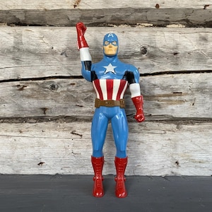 May include: A Captain America action figure, featuring a blue and red costume with a white star on the chest. The figure has a brown belt, red boots, and a raised fist. The background is a weathered wooden surface.