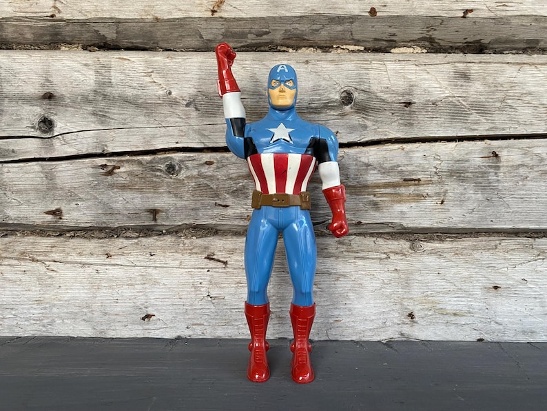 May include: A Captain America action figure, featuring a blue and red costume with a white star on the chest. The figure has a brown belt, red boots, and a raised fist. The background is a weathered wooden surface.