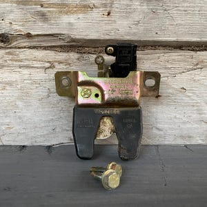 Puede incluir: Un conjunto de cierre de maletero de coche metálico con una base de plástico negro y dos pernos dorados. El cierre tiene un marco de metal plateado y dorado con marcas estampadas. El fondo es una superficie de madera envejecida.