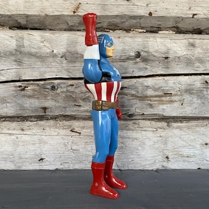 May include: A vintage Captain America action figure. The figure is in a side profile, with a blue and red costume, a white belt, and a red glove raised in the air. The figure is standing on a dark surface against a weathered wooden background.