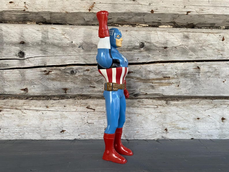 May include: A vintage Captain America action figure. The figure is in a side profile, with a blue and red costume, a white belt, and a red glove raised in the air. The figure is standing on a dark surface against a weathered wooden background.