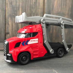 May include: A red toy truck with a gray loading mechanism. The truck has black wheels and the word "EXPRESS" on the side. The truck is on a wooden surface.