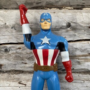 May include: A Captain America action figure in a classic pose. The figure is predominantly blue, with a white star on the chest, red and white stripes on the torso, and red gloves. It has a brown belt and a blue helmet with the letter "A".
