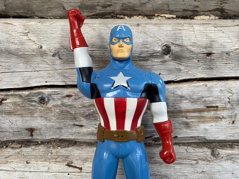 May include: A Captain America action figure in a classic pose. The figure is predominantly blue, with a white star on the chest, red and white stripes on the torso, and red gloves. It has a brown belt and a blue helmet with the letter "A".