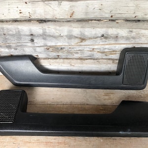 May include: Two black interior door armrests. Each armrest features a textured grip section and a smooth, curved design. The armrests are displayed against a weathered wooden backdrop, giving a vintage or rustic feel.