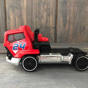 May include: A red and black toy truck with the number 24 in blue on the side. The truck has black wheels with silver rims and a silver undercarriage. The cab is red, and the trailer bed is black.