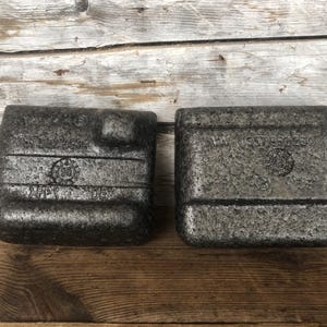 Puede incluir: Dos bloques rectangulares grises oscuros y texturizados con bordes redondeados. Cada bloque tiene texto en relieve y un emblema circular. Los bloques están sobre una superficie de madera.